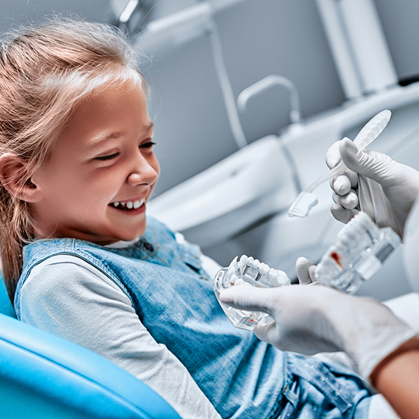 Young girl in dental chair grinning as dentist shows her a model of the teeth